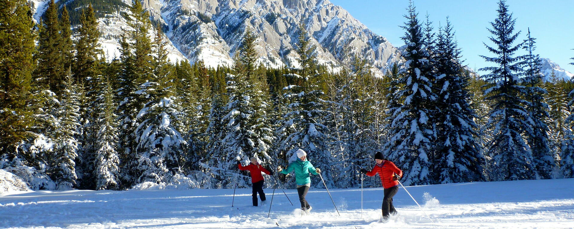 three skiers in open snowy field below rocky mountain