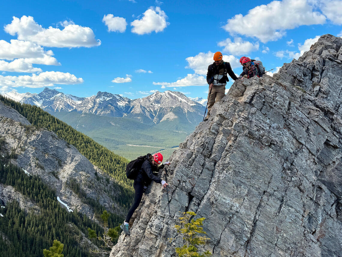 One person moves on rocky slope while two others look on in front of blue sky and mountain peaks behind
