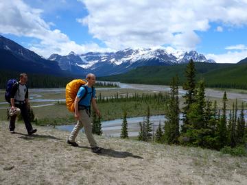 two backpackers cross along ridge overlooking braided river with mountain backdrop