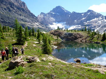 hikers walk narrow trail beside subalpine pond with mountains behind