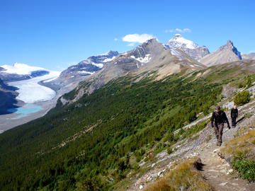 two hikers approach on trail with mountains and glacier behind