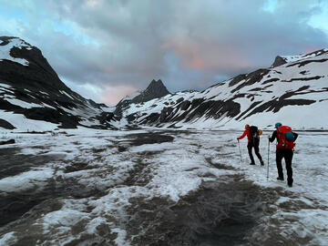 two people with backpacks walk across snowy floodplain toward mountains