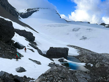 two people walk across steep snowy hillside above small tarns with mountain peak ahead