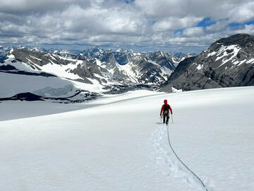 Person walks across broad snowy slope trailing rope and looking ahead toward mountains