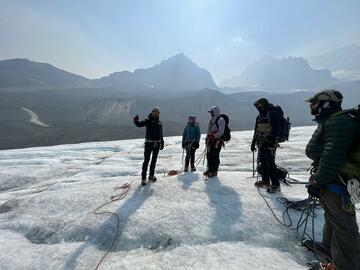 man gestures with four people onlooking. All stand on glacier geared up for mountain travel with ropes and harnesses.