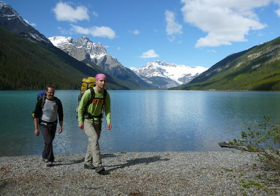 two men wearing backpacks walk into foreground with lake and mountains behind