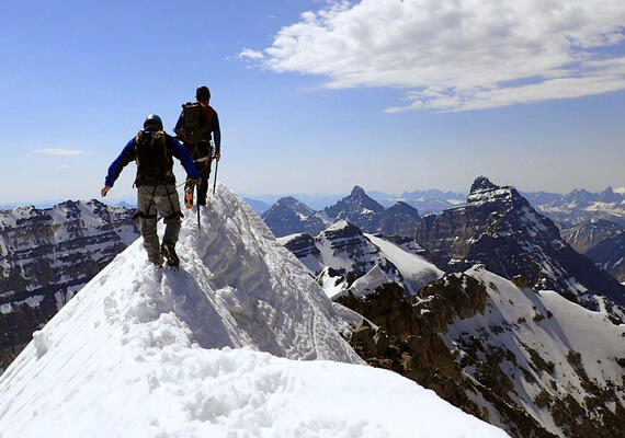 two people wearing helmets, harnesses, and backpacks walk along a narrow snow ridge while carrying ice axes in front of multiple rows of mountain peaks and ridges in background.